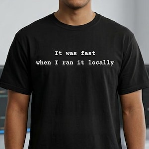 Può includere: T-shirt nera con la scritta bianca "It was fast when I ran it locally". La maglietta è indossata da una persona. Lo sfondo include una scrivania con monitor di computer.