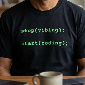 Puede incluir: Una camiseta negra con el texto "stop(vibing); start(coding);" en verde. La persona de la imagen está sentada en un escritorio con dos portátiles y una taza. El fondo incluye una estantería.