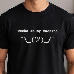 Può includere: T-shirt nero con la scritta "works on my machine" e un'emoticon stilizzata in bianco. Il testo e il disegno bianchi risaltano sul tessuto scuro. Un capo comodo e casual.
