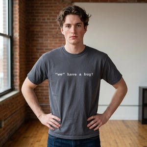 Peut inclure: Un t-shirt gris anthracite avec le texte blanc « we » have a bug ? Le t-shirt à manches courtes est fait d'une matière douce. La personne qui porte le t-shirt a les mains sur les hanches.