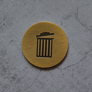 Puede incluir: Un objeto metálico redondo de color dorado con un icono de papelera negro. El diseño de la papelera está centrado en el disco, con una tapa y líneas verticales que representan el cuerpo del cubo. El fondo es una superficie gris texturizada.