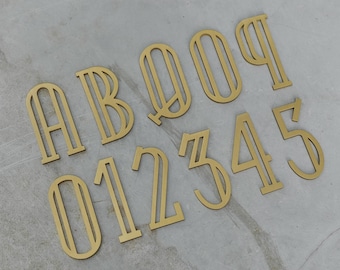 Könnte beinhalten: Eine Sammlung goldfarbener Metallbuchstaben und -zahlen, darunter A, B, 0, 1, 2, 3, 4 und 5, angeordnet auf einer hellgrauen Oberfläche. Die Zahlen und Buchstaben haben ein modernes, offenes Design.