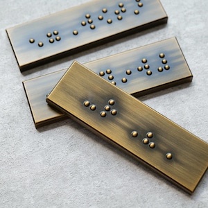May include: Three rectangular bronze-colored plaques with raised dots arranged in patterns, likely representing Braille. The plaques have a textured surface and a warm, metallic finish. The arrangement suggests a focus on tactile communication or design.
