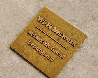 May include: A square, textured, golden-brown sign with the words "Wi-Fi Network" and "Belamaric Guest beourguest" in raised, gold lettering. A thin, gold line bisects the sign diagonally.