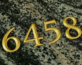 Könnte beinhalten: Goldfarbene Hausnummern aus Metall, 6, 4, 5 und 8, werden auf einer gesprenkelten grau-schwarzen Oberfläche angezeigt. Die Zahlen sind in einer klassischen Schriftart und scheinen dreidimensional zu sein.