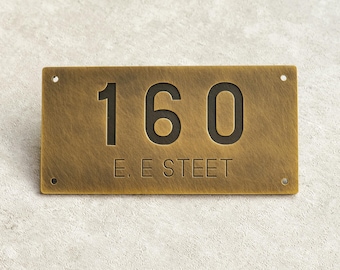 May include: A rectangular brass house number plaque with the number "160" in bold, black font. The text "E. E STREET" is below the number. The plaque has a textured surface and four mounting holes.