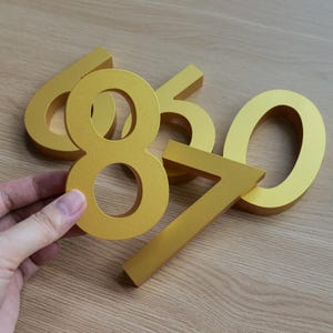 Könnte beinhalten: Goldfarbene Metallzahlen 6, 8, 5, 7 und 0. Die Zahl 8 wird von einer Hand gehalten. Die Zahlen sind in einer serifenlosen Schriftart und eignen sich für Hausnummern oder Beschilderungen.