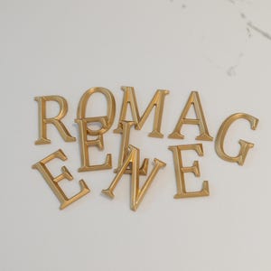 May include: Gold-colored metal letters spelling out the word "ROMANCE". The letters are in a classic serif font and are scattered on a white surface. The letters are three-dimensional and appear to be made of a shiny metal.