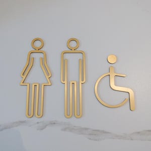 May include: Gold-colored restroom signs, including a female figure in a dress, a male figure, and a wheelchair symbol. The signs are made of a metallic material and are mounted on a white marble-patterned surface.