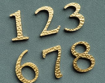 Könnte beinhalten: Nahaufnahme von goldenen Hausnummern aus Metall: 1, 2, 3, 6, 7 und 8. Jede Zahl hat eine gehämmerten Textur und wird vor einem gedämpften tealfarbenen Hintergrund angezeigt. Diese Nummern eignen sich für die Heimdekoration.