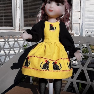 Puede incluir: Muñeca con un vestido amarillo bordado con gatos negros y un sombrero negro. El vestido tiene mangas negras y lazos rojos en los gatos. La muñeca tiene el pelo rosa y está sobre un soporte blanco.