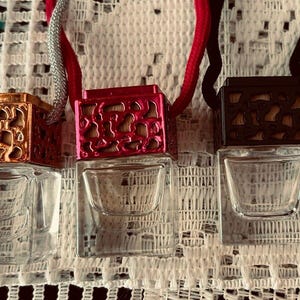 Può includere: Tre piccole bottiglie di vetro trasparente con coperchi quadrati e coperchi metallici decorativi. I coperchi sono dorati, rossi e neri, ciascuno con un motivo a intaglio unico. Le bottiglie sono appese a cordoncini argentati, rossi e neri. Sfondo bianco.