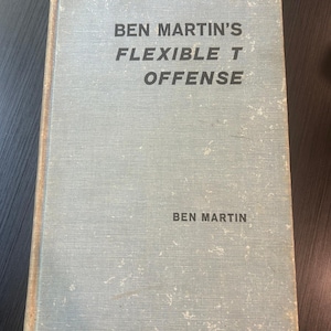 Könnte beinhalten: Ein gebundenes Buch mit einem hellblauen, strukturierten Stoffeinband. Der Titel "BEN MARTIN'S FLEXIBLE T OFFENSE" und der Autorenname "BEN MARTIN" sind in fetten, schwarzen, serifenlosen Großbuchstaben auf dem Buchdeckel aufgedruckt.