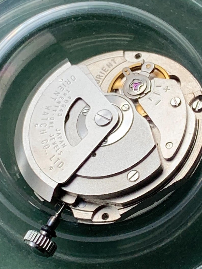 May include: Detailed view of a watch mechanism, revealing its complex internal workings. The silver components are marked with "Orient Watch Co. Japan" and "Twenty-One Jewels." A black stem with a textured crown is present. The background is a translucent green.