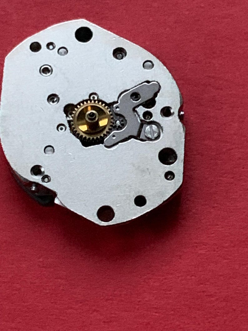 May include: Close-up of a watch movement component. The silver-colored piece has multiple holes and a gold-colored gear in the center. A small, silver-colored lever is also visible. The background is a solid red color.