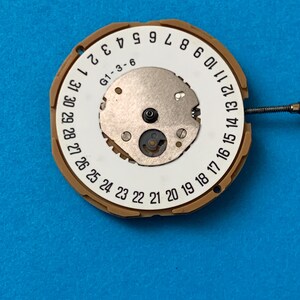 May include: A watch movement with a white dial featuring black numerals from 1 to 31, indicating the days of the month. The dial has a central metal component and is surrounded by a tan-colored ring. The text "G1-3-6" is visible.