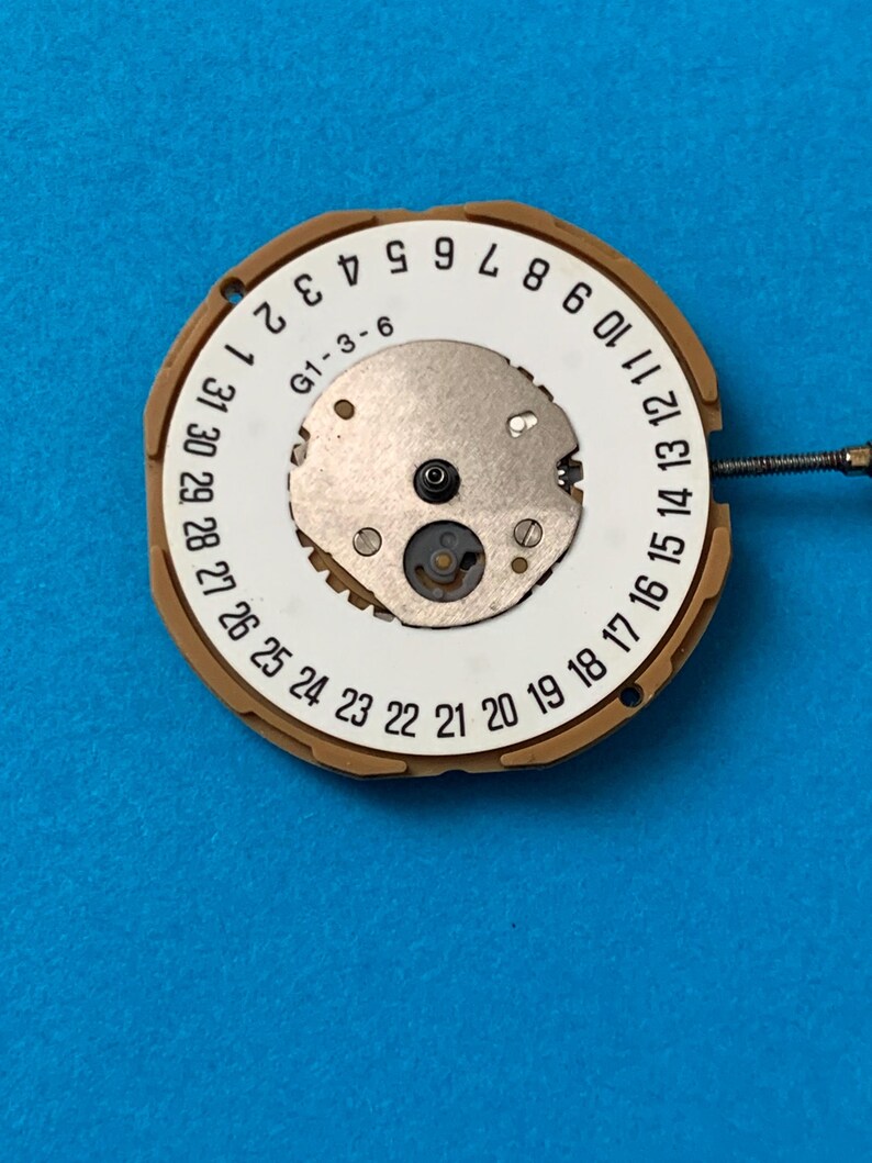 May include: A watch movement with a white dial featuring black numerals from 1 to 31, indicating the days of the month. The dial has a central metal component and is surrounded by a tan-colored ring. The text "G1-3-6" is visible.