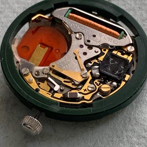 May include: Close-up of a Citizen watch movement. The watch mechanism features gold-colored gears, a copper-colored coil, and an orange battery component. The movement is set within a dark green circular frame, with a silver crown visible.