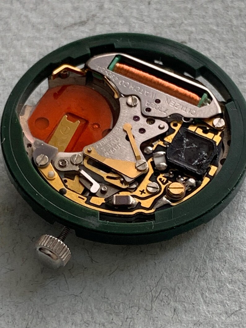 May include: Close-up of a Citizen watch movement. The watch mechanism features gold-colored gears, a copper-colored coil, and an orange battery component. The movement is set within a dark green circular frame, with a silver crown visible.