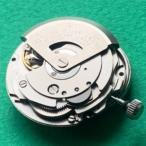 May include: Close-up of a silver Orient Twenty-One Jewels watch movement. The intricate mechanism features gears, screws, and a winding stem. The watch movement is set against a green background.