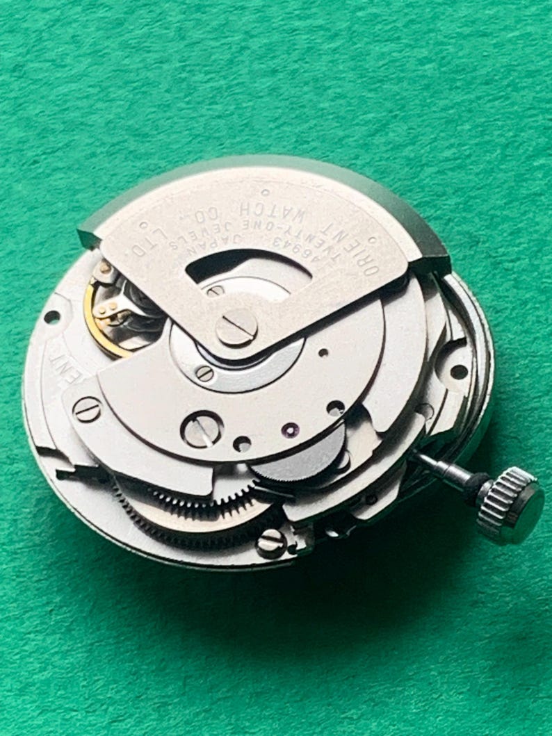 May include: Close-up of a silver Orient Twenty-One Jewels watch movement. The intricate mechanism features gears, screws, and a winding stem. The watch movement is set against a green background.