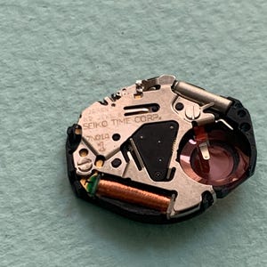 May include: A close-up of a watch movement with the text "SEIKO TIME CORP." and "JAPAN" visible. The movement has a silver-coloured metal plate, a black plastic base, and copper-coloured coils. A small red component is also visible.