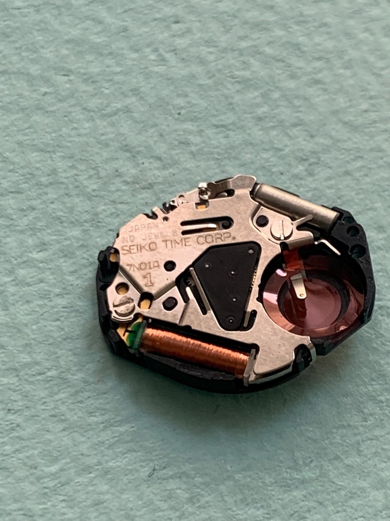 May include: A close-up of a watch movement with the text "SEIKO TIME CORP." and "JAPAN" visible. The movement has a silver-coloured metal plate, a black plastic base, and copper-coloured coils. A small red component is also visible.