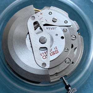 May include: A close-up of a Seiko 21 Jewels watch movement. The silver mechanism has the text "SEIKO 21 JEWELS" and "JAPAN". The watch movement is set in a clear plastic container.