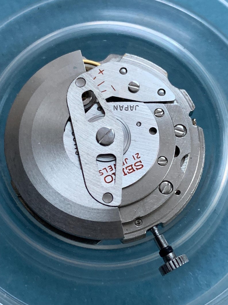 May include: A close-up of a Seiko 21 Jewels watch movement. The silver mechanism has the text "SEIKO 21 JEWELS" and "JAPAN". The watch movement is set in a clear plastic container.