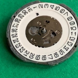 May include: Close-up of a watch movement component. The component features a white circular dial with black numbers indicating the days of the month, from 1 to 31. The dial is surrounded by a gray ring. The center reveals the watch's internal mechanisms.