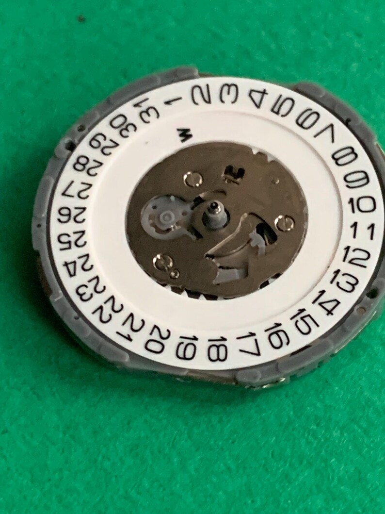 May include: Close-up of a watch movement component. The component features a white circular dial with black numbers indicating the days of the month, from 1 to 31. The dial is surrounded by a gray ring. The center reveals the watch's internal mechanisms.
