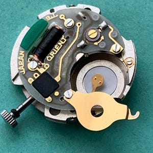 May include: Close-up of a watch movement with the word "ORIENT" visible. The movement features gold-colored components, a black coil, and a green component labeled "52950". The word "JAPAN" is also visible.