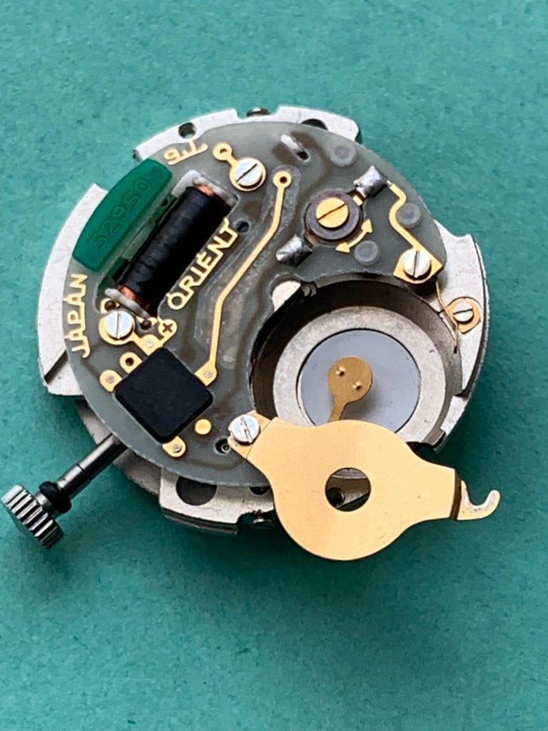 May include: Close-up of a watch movement with the word "ORIENT" visible. The movement features gold-colored components, a black coil, and a green component labeled "52950". The word "JAPAN" is also visible.