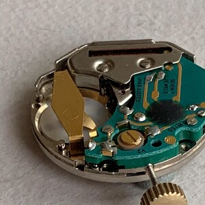 Puede incluir: Primer plano de un mecanismo de reloj con una placa de circuito verde, componentes dorados y una carcasa de metal plateado. El mecanismo del reloj incluye una corona y el texto "UNADJUSTED" y "SEVEN 7 JEWELS".