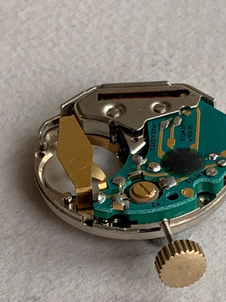 Puede incluir: Primer plano de un mecanismo de reloj con una placa de circuito verde, componentes dorados y una carcasa de metal plateado. El mecanismo del reloj incluye una corona y el texto "UNADJUSTED" y "SEVEN 7 JEWELS".