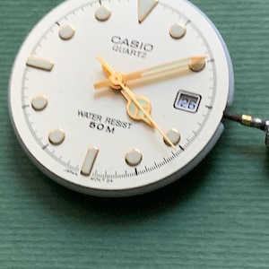 K&ouml;nnte beinhalten: Ein Casio Quartz Zifferblatt mit cremefarbenem Zifferblatt, goldenen Zeigern und Markierungen. Das Zifferblatt zeigt "CASIO QUARTZ" und "WATER RESIST 50M". Das Datum wird als "26" angezeigt.