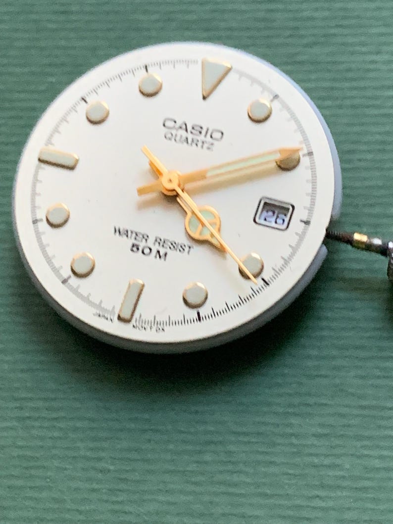 K&ouml;nnte beinhalten: Ein Casio Quartz Zifferblatt mit cremefarbenem Zifferblatt, goldenen Zeigern und Markierungen. Das Zifferblatt zeigt "CASIO QUARTZ" und "WATER RESIST 50M". Das Datum wird als "26" angezeigt.