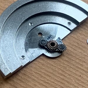 Pu&ograve; includere: Primo piano di componenti di orologi, tra cui una piastra semicircolare color argento con tre piccole viti e una serie di minuscoli ingranaggi. Gli ingranaggi sono argentati e bronzo, e sono posizionati sulla piastra.