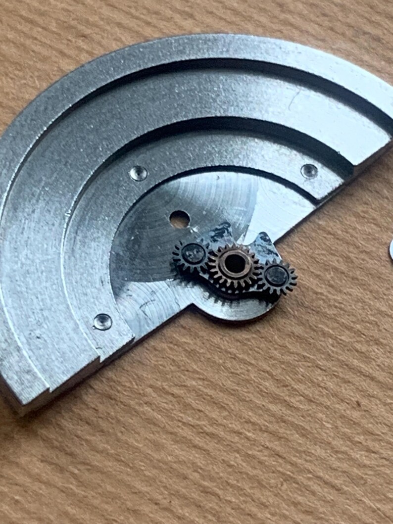 Pu&ograve; includere: Primo piano di componenti di orologi, tra cui una piastra semicircolare color argento con tre piccole viti e una serie di minuscoli ingranaggi. Gli ingranaggi sono argentati e bronzo, e sono posizionati sulla piastra.
