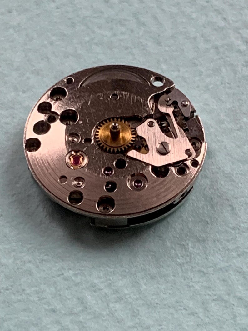 May include: Close-up of a silver watch movement with a gold gear and a red jewel. The movement is on a white surface.