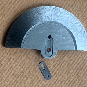Pu&ograve; includere: Primo piano di componenti di orologi. Il pezzo principale &egrave; un componente semicircolare in metallo spazzolato con un foro centrale allungato e un piccolo ingranaggio. &Egrave; presente anche un pezzo pi&ugrave; piccolo, rettangolare, a forma di buco della serratura.