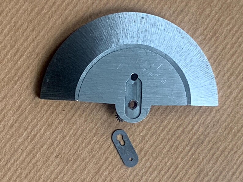 Pu&ograve; includere: Primo piano di componenti di orologi. Il pezzo principale &egrave; un componente semicircolare in metallo spazzolato con un foro centrale allungato e un piccolo ingranaggio. &Egrave; presente anche un pezzo pi&ugrave; piccolo, rettangolare, a forma di buco della serratura.