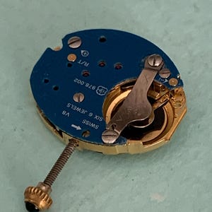 May include: A close-up of a watch movement with a blue circuit board and gold-colored components. The watch movement has a screw with a gold crown and a silver lever. The text "SWISS SIX 6 JEWELS" is visible.