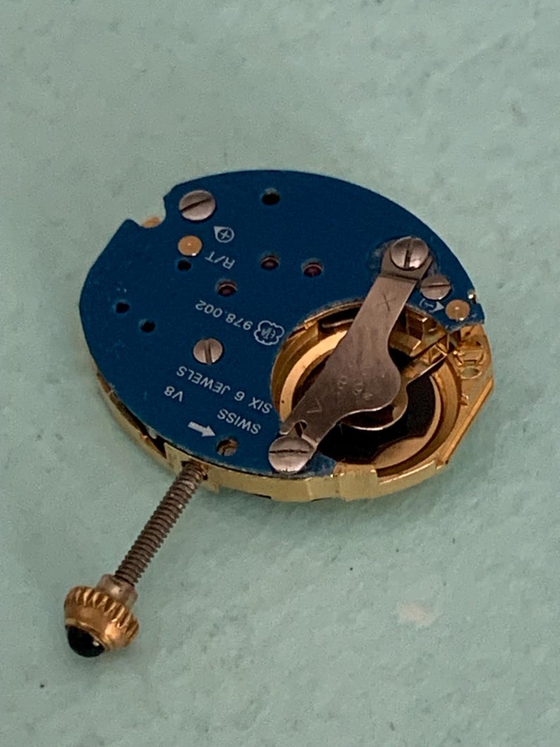 May include: A close-up of a watch movement with a blue circuit board and gold-colored components. The watch movement has a screw with a gold crown and a silver lever. The text "SWISS SIX 6 JEWELS" is visible.