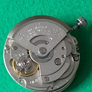 May include: Close-up of a watch movement with silver-colored gears and components. The watch mechanism has a small crown and the text "JAPAN SEVEN JEWELS CO. LTD." is visible. The background is a solid green.