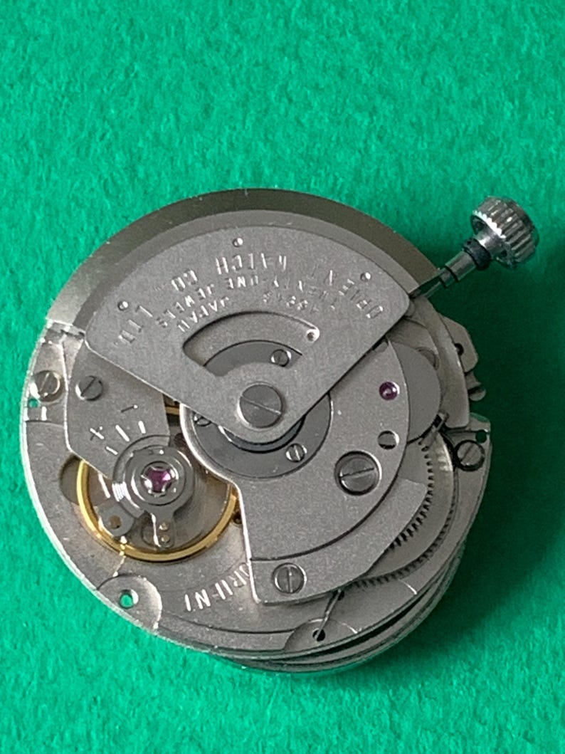 May include: Close-up of a watch movement with silver-colored gears and components. The watch mechanism has a small crown and the text "JAPAN SEVEN JEWELS CO. LTD." is visible. The background is a solid green.