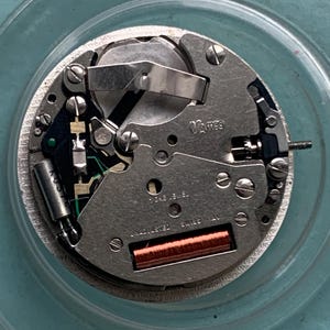 Op de afbeelding: Close-up van een uurwerk met zilverkleurige metalen componenten, schroeven en een koperkleurige spoel. De tekst "Vames" en "Swiss Made" zijn zichtbaar. Het uurwerk ligt op een lichtblauwe ondergrond.