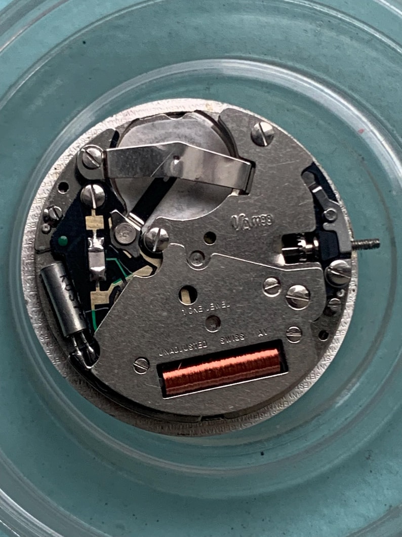 Op de afbeelding: Close-up van een uurwerk met zilverkleurige metalen componenten, schroeven en een koperkleurige spoel. De tekst "Vames" en "Swiss Made" zijn zichtbaar. Het uurwerk ligt op een lichtblauwe ondergrond.