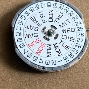 May include: A watch calendar dial with a white face and a gray metal frame. The days of the week are printed in black and red, with the numbers 1-31 around the edge. The dial is a circular shape.