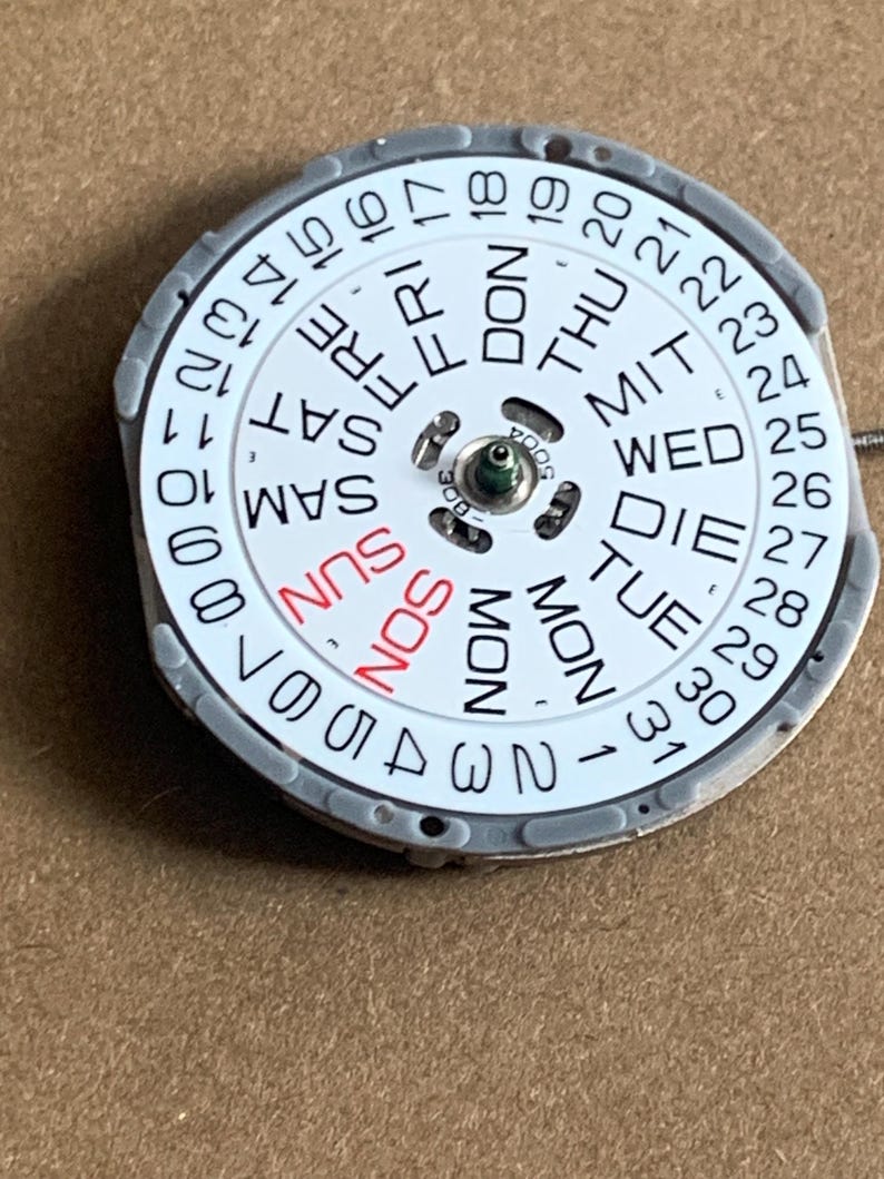 May include: A watch calendar dial with a white face and a gray metal frame. The days of the week are printed in black and red, with the numbers 1-31 around the edge. The dial is a circular shape.